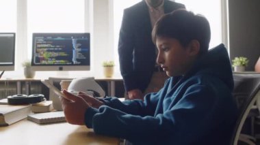 Orta boy Hintli bir çocuğun okulda bilgisayar bilimi dersinde otururken, AR kulaklığı için tablet üzerinde uygulama geliştirirken, Orta Doğulu bir erkek öğretmenin yardım teklif etmek için yürüdüğü bir fotoğraf.
