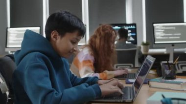 Okulda programlama dersinde oturan, dizüstü bilgisayardaki kodlama projesi üzerinde çalışan, kameraya bakıp gülümseyen ve sınıfta gezinen Hintli bir çocuğun orta boy portresi.