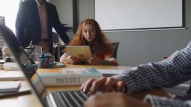 Programlama dersinde çalışan genç öğrencilerin orta boy fotoğrafı, kızıl saçlı genç kıza yaklaşan erkek öğretmen, tablet üzerinde mobil uygulama üzerinde çalışıyor ve ilerlemeyi kontrol ediyor.