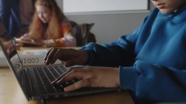 Programlama dersinde oturan, dizüstü bilgisayara kod yazmaya, klavyeye yazmaya, ekrana dikkatle bakmaya odaklanmış, mavi eşofmanlı 12 yaşındaki inek öğrencinin orta boy yakın çekimi.