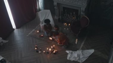 Yerde yanan mumların arasında oturan ve karanlık köşkte yaşayan karanlık güçleri çağıran gizemli kitaplardan büyülü sözler okuyan üç gencin yukarıdan çekimleri.