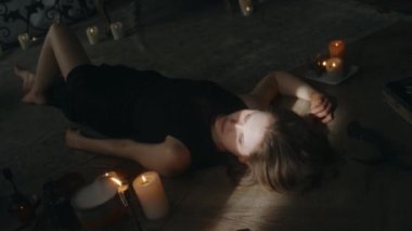 Beyaz, siyah elbiseli genç bir cadının, yanan mumların arasında, ahşap zeminde yatarken ve evde karanlık ayinler yaparken gözlerini açarken boy açışı.