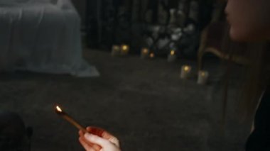 Yanan mumların arasında oturan ve palo santo ağacını yakan genç cadının yan yatay görüntüsü. Negatif enerjiden evi temizlemek ve karanlık ayinlere yer açmak için onu üflüyor.