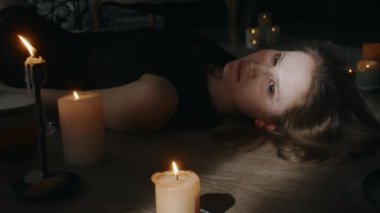 Şeytan tarafından ele geçirilmiş siyah elbiseli beyaz bir kadının ahşap bir zeminde yanan mumların arasında uyandığı bir resim.