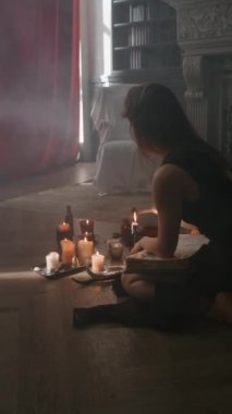 Dikey çekimde, siyah elbiseli gizemli bir cadının mum yakarken, okült bir kitapla ve palo santo ağacını yakarken karanlık seremoniye hazırlanırken.