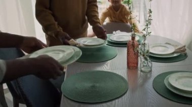 Yemek masasına tabak ve peçeteleri koyan tanınmayan Afrikalı Amerikalı erkek aile üyelerinin orta boy yakın çekimleri ve orta kesimi.