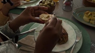 Örgü ören, kızarmış ekmeğe tereyağı veya ezme süren genç Afrikalı Amerikalı kadının elleri omuz üstü, kızarmış tavuk ve sebzelerle kaplı tabakta, aile veya arkadaşlarıyla akşam yemeğinde.
