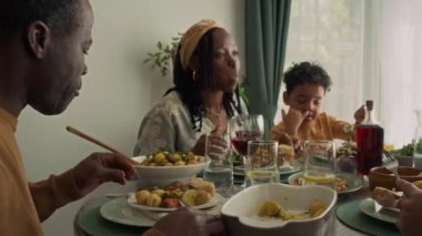Orta boy, örgülü, Afrika kökenli Amerikalı bir kadın ve 6 yaşındaki oğlu tatilde aile partisinde masada oturmuş, bayram yemeğinin tadını çıkarıyor, kızarmış tavuk yiyor ve şarap içiyor.