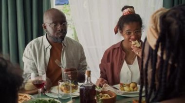 Orta boy Afrikalı Amerikalı bir kız ve sakallı bir baba aile partisinde masada oturmuş leziz ev yapımı rosto yiyor, tost çiğniyor ve sohbet ederken