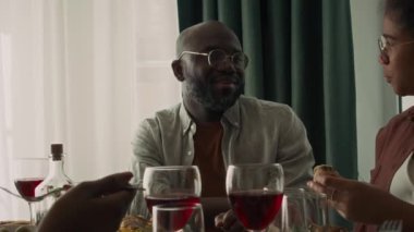 Orta boy, gözlüklü Afro-Amerikan bir adamın, kızı, babası ve akrabalarıyla yemek masasında oturmuş lezzetli ev yemeği yerken ve tatilin tadını çıkarırken görüntüsü.