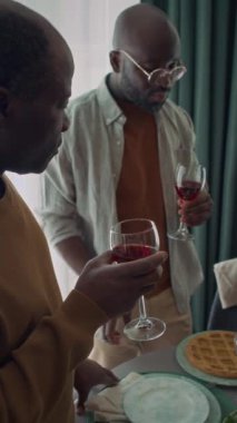 Orta yaşlı bir baba ve Afrikalı genç bir adamın oturma odasında gözlüklü bir şekilde kırmızı ev yapımı şarap içerken aile toplantısı sırasında lezzet ve buketi tartışırken dikey bir fotoğrafı.