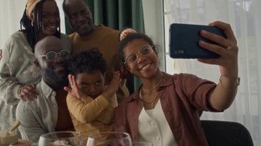Neşeli siyahi ailenin orta boy yakın çekimi - karısı, kocası, kızı, oğlu, büyükbabası, akşam yemeğinde eğlenceli selfie 'ler için poz vermeleri, sonra da akıllı telefondan fotoğrafa bakması, işaret etmesi ve gülmesi