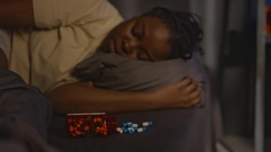 Açık plastik kavanozun orta boy görüntüsü. Yatağında sakinleştirici haplar var. Genç siyahi kadın geceleri arka planda uyuyor.