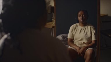 Genç siyahi bir kadının yatak odasındaki aynanın önünde otururken ve yansımasına bakarken omuz üstünden çekilen fotoğrafı. Kendinden kopuk hissediyordu.