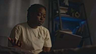 Düşük açılı genç Afro-Amerikan kadın ev kıyafetleri içinde yatağında oturuyor ve dizüstü bilgisayarında psikiyatrist ile internetteki oturumunu bitiriyor uykusuz bir geceden sonra yorgun görünüyor, travma sonrası stres bozukluğu yaşıyor.