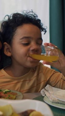 5 yaşındaki Afro-Amerikan çocuğun aile yemeği partisinde masada oturup bir bardak portakal suyu içerken ve annesinin omzunu sıvazlarken orta boy dikey çekimi.