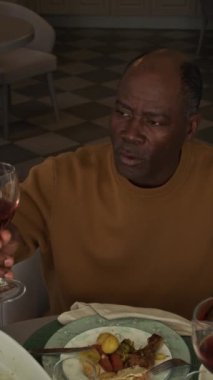 Orta yaşlı Afrikalı Amerikalı bir adamın kadehini şarapla kaldırırken, arkadaşlarıyla ya da aile üyeleriyle tokuştururken ve ev partisinde yemek yerken orta boy dikey yavaş çekimleri.