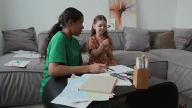 Orta boy Afro-Amerikan bebek bakıcısının boş zamanlarında evde Kafkasyalı anaokulu kızıyla etkileşim halindeyken, renkli resimler çizerken ve konuşurken kahve masasının başında otururken.