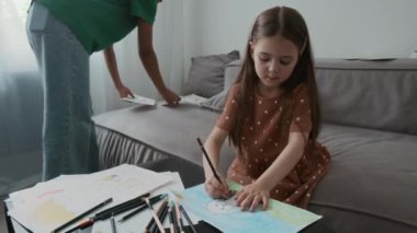 Beyaz kız renkli kalemlerle kağıt üzerine resim çizerken Afrikalı Amerikalı genç bebek bakıcısının koltuktan alfabe kartlarını kaldırdığı orta boy bir fotoğraf.