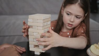 Kafkasyalı anaokulu öğrencisi kızın evde arkadaşıyla masa oyunu oynaması, orta seviyeden tahta bloğu alıp en üste koyması.