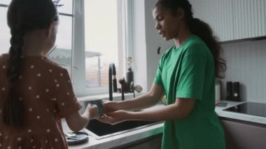 Afro-Amerikalı genç bebek bakıcısı Kafkas anaokulu kızına bulaşıkları yıkama ve silme dersi veriyor, minimalist apartmanda birlikte ev işi yapıyorlar.