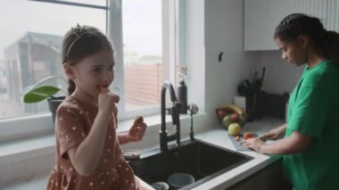 Orta boy beyaz bir kız. Lavabonun yanındaki yemek masasında oturup havuç yerken Afro-Amerikan genç dadısıyla yemek pişiriyor.