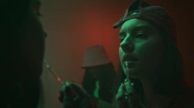 Kısa saçlı ve şapkalı genç bir kadının karanlık neon ışıklı tuvaletlerde dudak parlatıcısı sürerken çekilmiş yakın plan ayna görüntüsü.