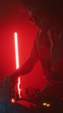 Süslü gömlekli ve güneş gözlüklü profesyonel hipster DJ 'in dikey yan görüntüsü. Düğmeleri ve kaydırakları çevirirken mikser kontrolörünü kullanıyor ve kırmızı neon ışıklı gece kulübünde halka EDM çalıyor.