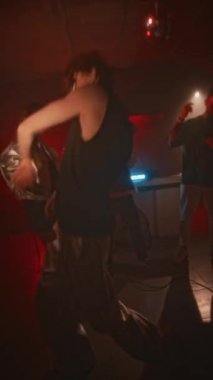 Tekno çılgın güneş gözlüklü, atlet tişörtlü, gümüş krom pantolonlu, gece kulübünde kırmızı neon ışıklı ve sürekli yanıp sönen neşeli dans müzikleri yapan yüzsüz adamın dikey pozu.