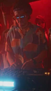 Havacı gözlüklü ve havalı tişörtlü havalı hippi adamın dikey el kamerası görüntüsü. DJ ses mikser kontrolörüyle eğlenceli bir kalabalık neon ışıklı gece kulübü partisinde EDM çalıyor.