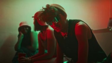 Bereli kendinden emin bir partinin orta boy portresi ve aynalı tekno güneş gözlüğü. Dumanlı neon ışıklı gece kulübü odasında oturup kameraya bakarken şişeden bira içiyorlar.