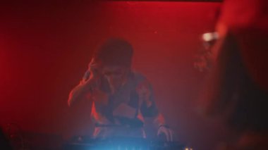 Süslü tişörtlü, gözlüklü hippi adamın ses mikserinde DJ 'lik yaptığı ve gece kulübünde kırmızı neon ışıklı dans eden kalabalığa DJlik yaptığı el kamerası görüntüsü.