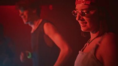 Retro y2k kıyafetleri içinde genç bir bayan ve adamın kalabalık içinde coşup kırmızı neon ışıklı gece kulübündeki elektronik dans müziğine geçişlerinin yan görüntüsü.