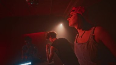 Kırmızı neon ışıklı gece kulübündeki retro partisinde elektronik dans müziğine gevşek hareketler yapan kot bluzlu, kep ve oval güneş gözlüklü, kendine güvenen genç bayanın el kamerası görüntüsü.
