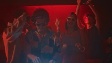 DJ konsolunda iyimser müzik çalan havalı hippi adamın orta boy el kamerası görüntüsü ve arkasında dans eden gençlerle dolu neon ışıklı ve yanıp sönen bir kulüpte.