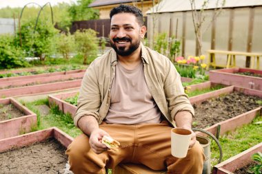 İş kıyafetleri içinde neşeli bahçıvan elinde sandviç ve kahve tutarken kameranın önünde yeşil fidelerle çiçek tarlalarına karşı oturuyor.