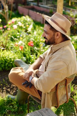 Çiçekler ve yeşil bitkilerle çiçeklerin önünde otururken iş kıyafetleri ve şapkalı, sakallı, olgun bir erkek bahçıvan ileriye bakıyor.