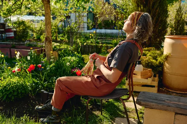 stock image Side view of mature female gardener in grey t-shirt, brown overalls and headband sitting on chair in the garden and enjoying sunlight