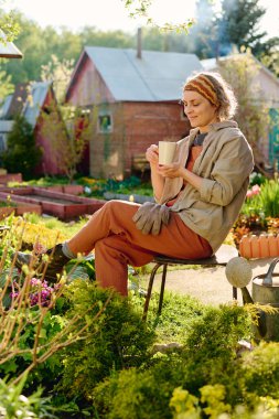 İş kıyafetleri içinde gülümseyen kadın bahçıvan yeşil bitkilerin arasında otururken elinde sıcak çay veya aromatik kahveyle fincana bakıyor.