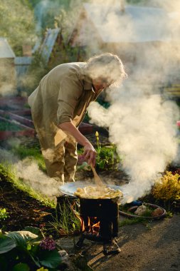 Yetişkin kadın bahçıvan iş kıyafetleri içinde sıcak tava ve duman üzerinde eğilerek bahçede yetişen doğranmış patatesleri karıştırıyor.