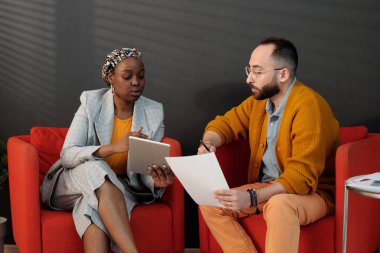 İki meslektaş kırmızı sandalyelerde oturuyor, proje detaylarını tartışıyor, tablet ve belge kullanıyor, işbirliği yapıyor ve ofiste görüşleri paylaşıyor