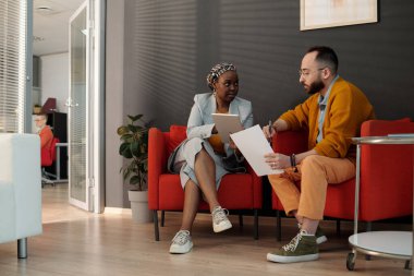 Modern bir ofis salonundaki iş arkadaşları, siyahi iş kadını tablet ve beyaz adamın elinde belgeler tutarken, her ikisi de tartışmalarına odaklanıyor.