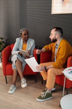 Modern ofis salonundaki meslektaşlar iş tartışıyor, siyahi iş kadını tablet kullanıyor ve beyaz adam konuşma sırasında belge tutuyor.