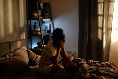 Uykusuz genç kadın gecenin bir yarısı yatakta otururken şakaklarına dokunuyor ve uykusuzluk ya da travma sonrası stres bozukluğu çekiyor.