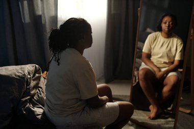 Karanlık bir yatak odasında aynanın önünde oturan ve travma sonrası sendromu geçiren depresyonlu genç bir kadın.