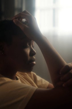 Kafasına dokunan ve gözlerini beyaz perdelerle pencereye kapatan Afrikalı Amerikalı genç bir kadının yan görüntüsü