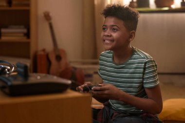 Kıvırcık saçlı Afro-Amerikalı genç çocuk, rahat bir odada video oyununa odaklanmış, yerde oturuyor ve oyun kumandasını tutarken ekranı seyrediyor.