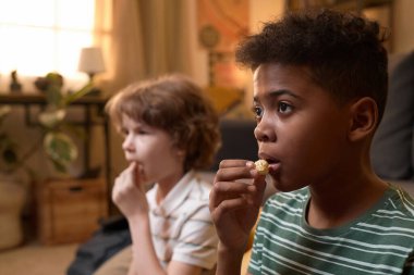 İki farklı çocuk oturmuş patlamış mısır yerken aynı zamanda eski moda oturma odasında yüzünde odaklanmış ifadeyle televizyon izliyorlar.