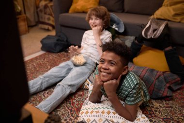 İki beyaz ve Afrikalı Amerikalı çocuk yerde yatıp ellerinde patlamış mısırla komik bir TV şovu izleyip rahat bir şekilde eski moda oturma odasında vakit geçiriyorlar.
