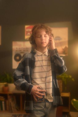 Klasik kaset çalar tutan, kulaklıklı ve kapalı gözlerle müzik dinleyen, duvarlarında posterlerle rahat bir odada duran genç bir çocuk.